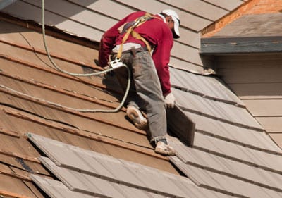 A roofer in a red shirt harnessed to a rope while laying down shingles.