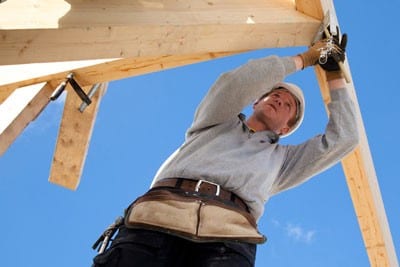 A construction worker in a gray sweater fixing the wooden frame of a house.